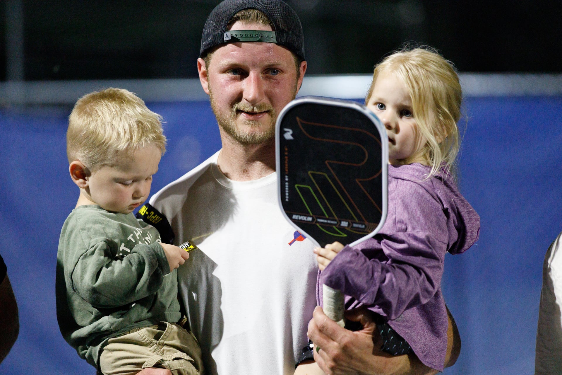 Man holding two children, one with a paddle, on a sports court.
