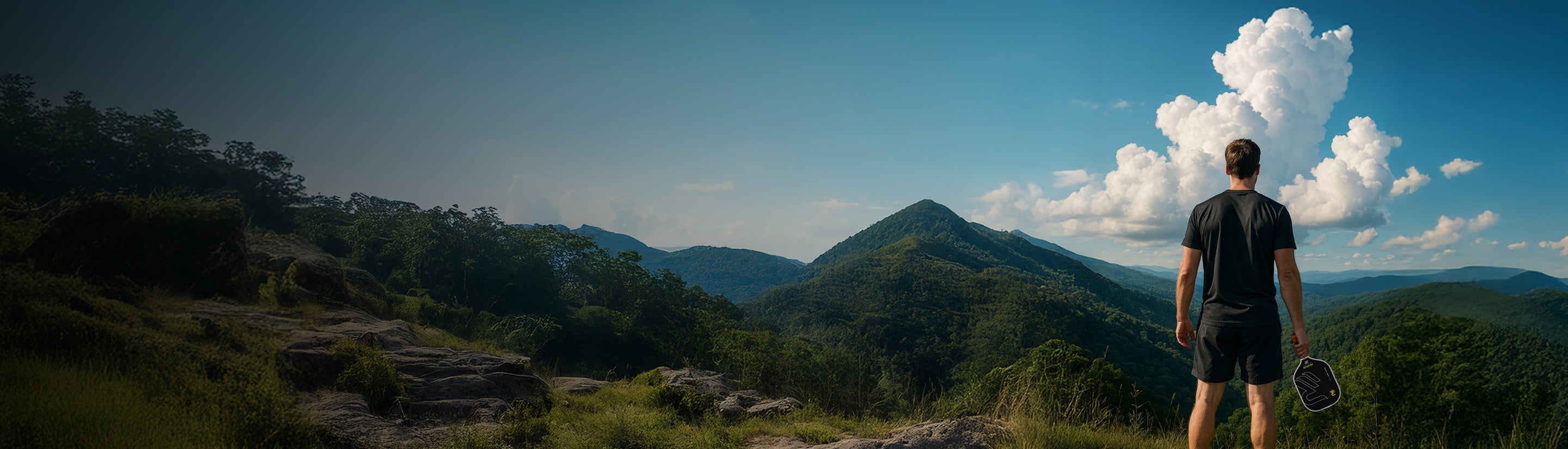 Person standing on a mountain top with a Revolin pickleball paddle and a scenic view of clouds and mountains.