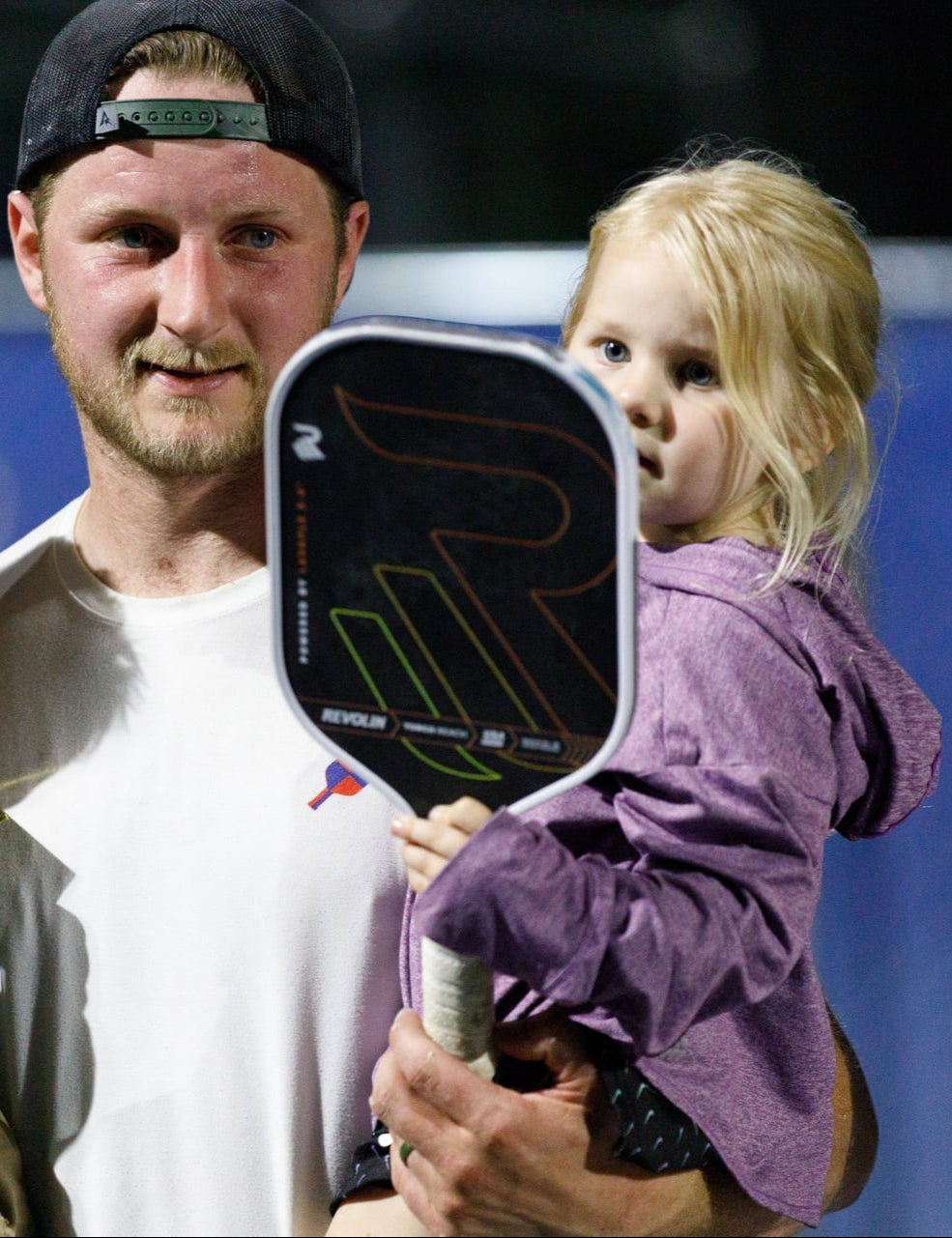 Man holding two children, one with a paddle, on a sports court.