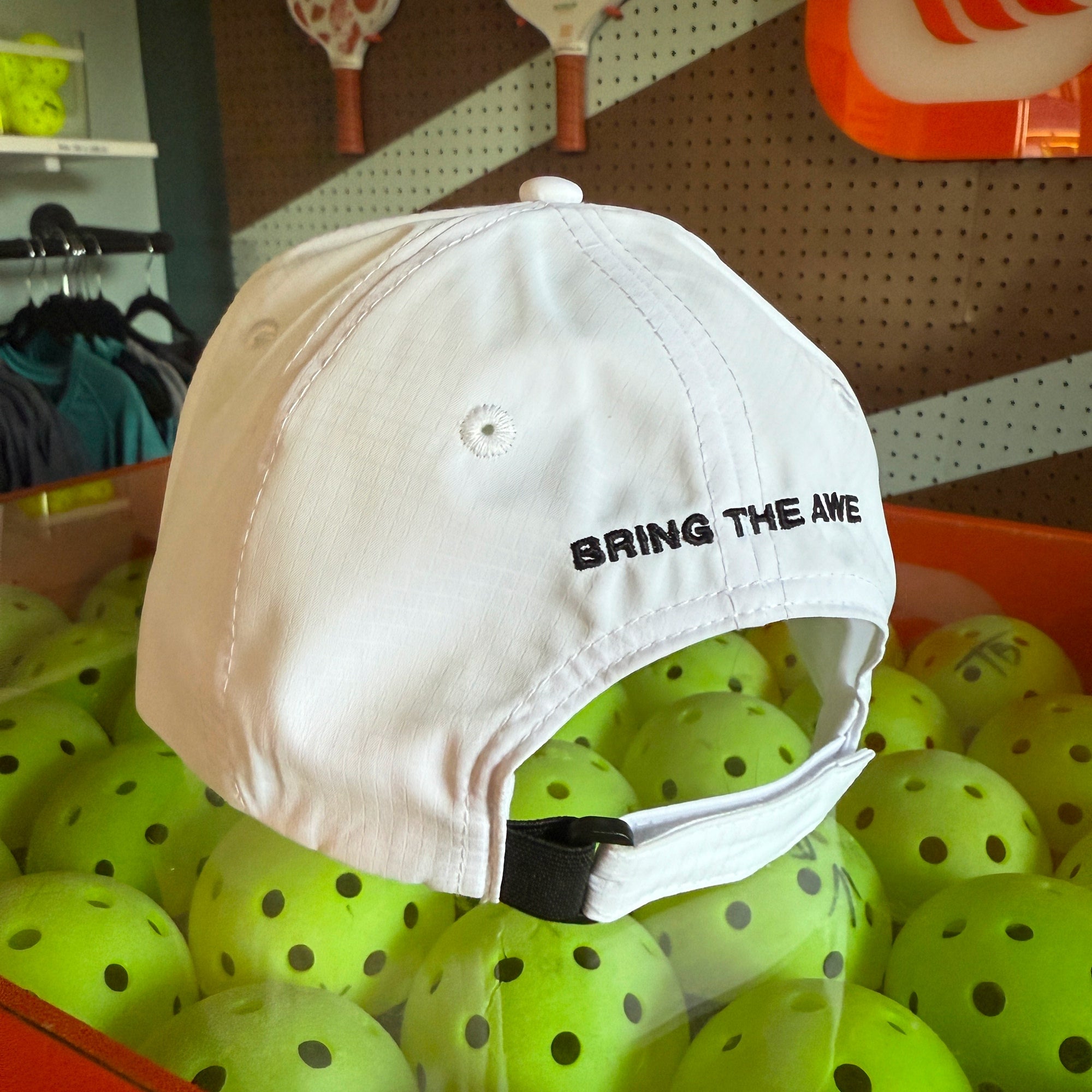 White cap with 'BRING THE AWE' text on a pile of pickleball balls in a store setting
